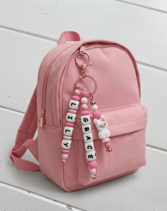 An elegant close-up of a personalized name charm held in hands against a soft, floral background. A perfect baby shower gift idea that grows with the child, transitioning from a pram accessory to a school backpack charm.
