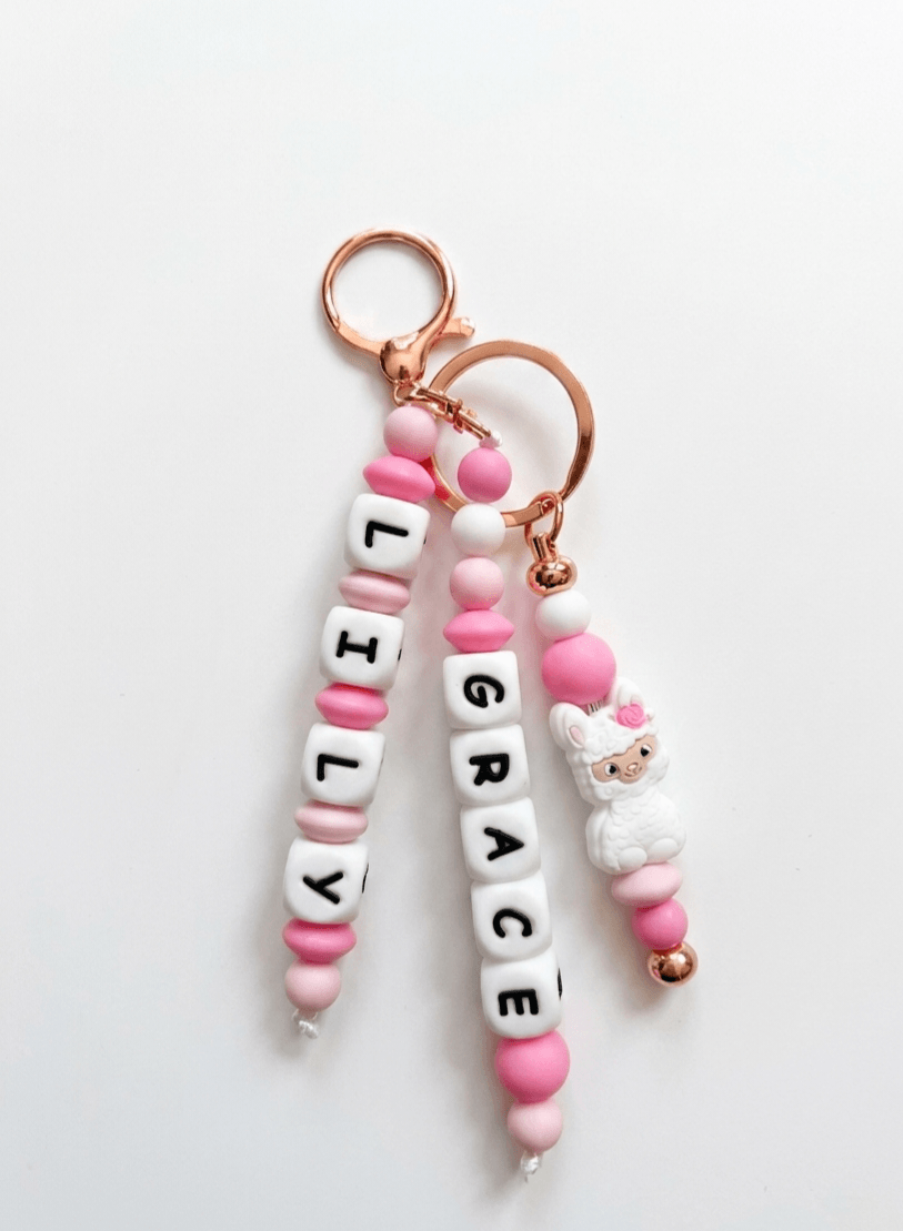 Handcrafted, personalized charm made from BPA-free soft silicone in shades of pink and white. It features three strands: two with name beads and one with a cute white llama figurine with a pink rose, all attached to an elegant rose gold clasp.