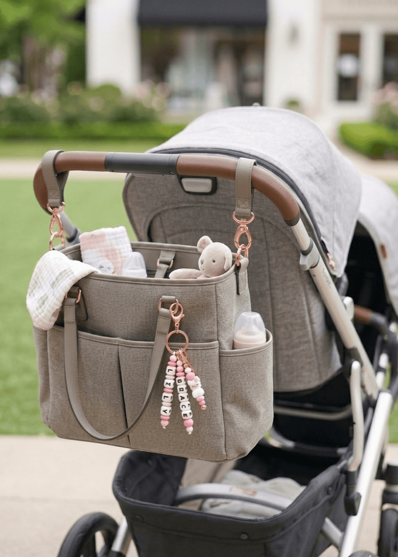 An elegant close-up of a personalized name charm held in hands against a soft, floral background. A perfect baby shower gift idea that grows with the child, transitioning from a pram accessory to a school backpack charm.