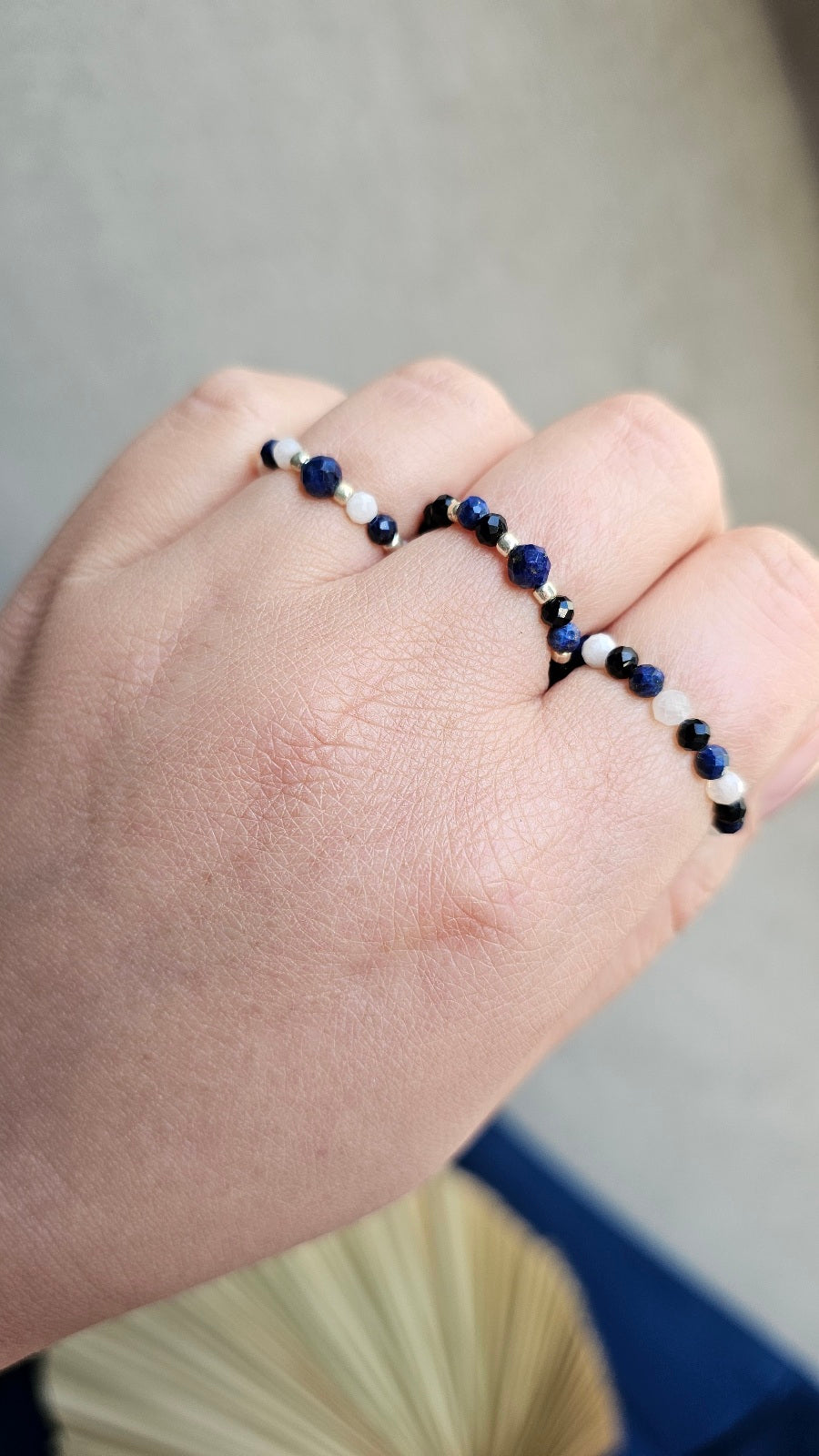 Three handmade elastic gemstone rings with Lapis Lazuli, Black Tourmaline and Moonstone beads, featuring gold and silver accents on a minimalist background.