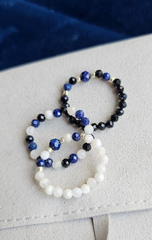 Three handmade elastic gemstone rings with Lapis Lazuli, Black Tourmaline and Moonstone beads, featuring gold and silver accents on a minimalist background.