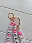 An elegant close-up of a personalized name charm held in hands against a soft, floral background. A perfect baby shower gift idea that grows with the child, transitioning from a pram accessory to a school backpack charm.