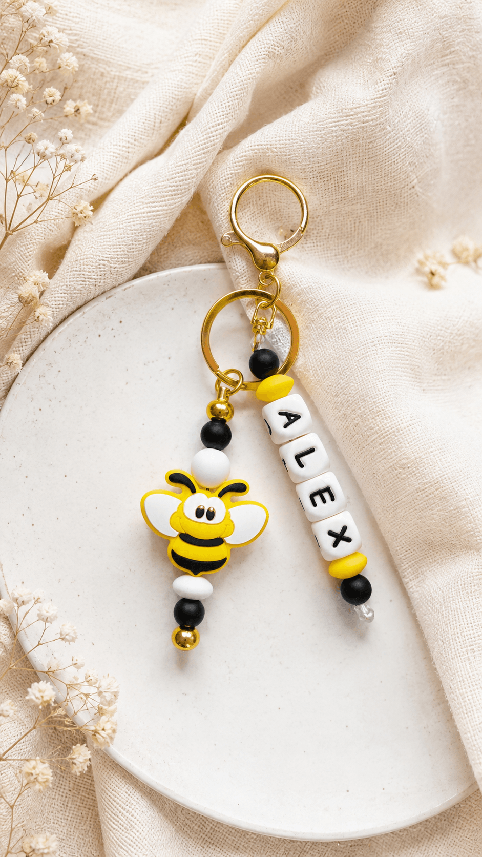 Close-up of a handmade personalised bee bag charm with name beads, gold hardware and yellow, black and white silicone beads on a soft cream background. A cute personalised gift for children, school bags and backpacks, handmade in Ireland.