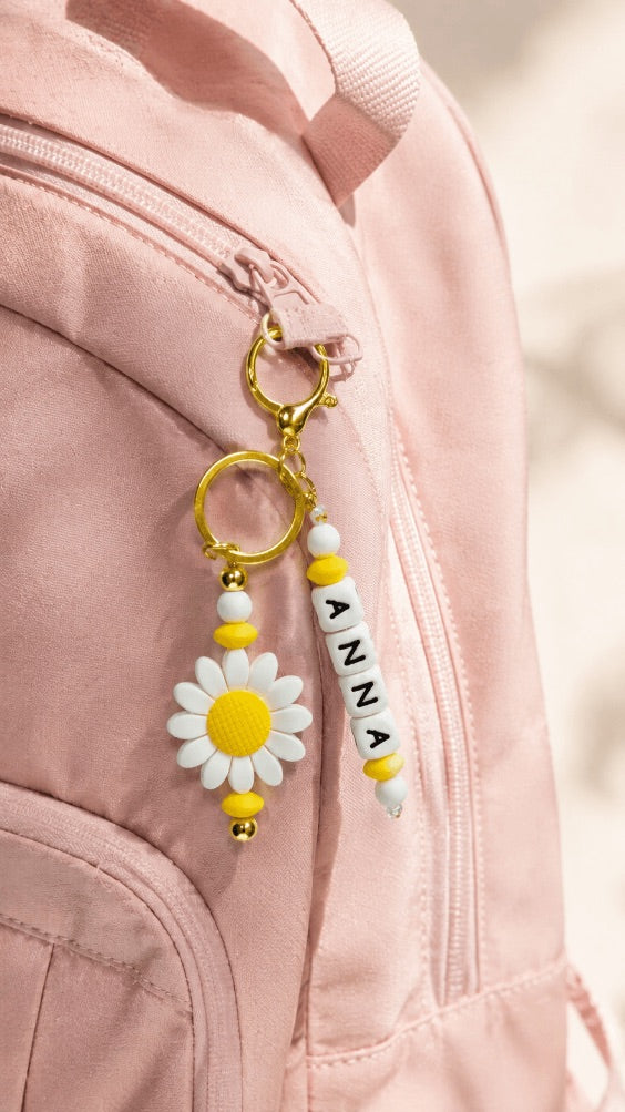 Personalised daisy bag charm with custom ANNA name, white and yellow silicone beads and gold-tone hardware attached to a beige and pink sports bag.