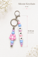 Elegant silicone bag charm with purple, blue and soft pink beads and silver-tone hardware on a soft cream background.
