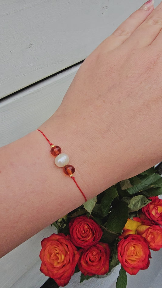 Close-up of a delicate red thread bracelet featuring a piece of golden Baltic amber and a natural freshwater pearl. The bracelet is gently tied around a wrist, symbolising luck, protection, and feminine energy. Soft natural light enhances the warm tones of the stones.
